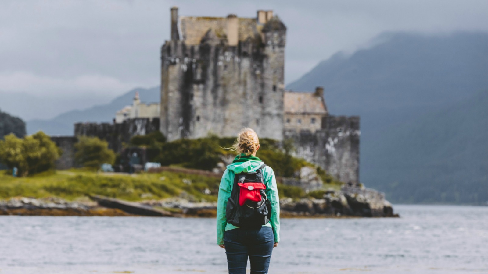 Young woman with a backpack standing in front of Eilean Donan Castle, framed by the surrounding mountains