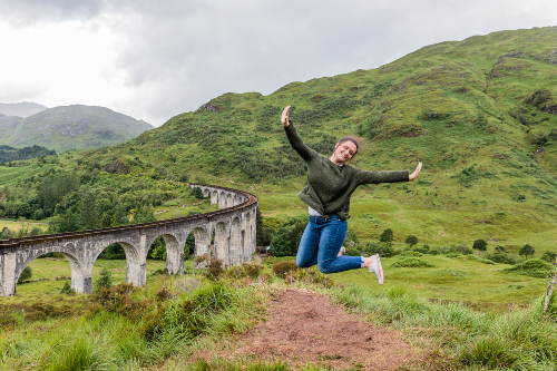 Young woman joyfully leaping into the air with the Glenfinnan Viaduct and Highland hills behind her