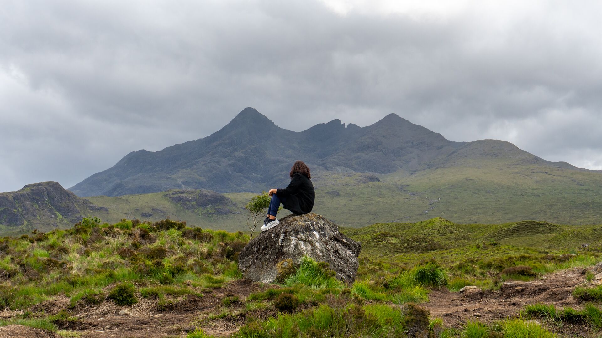 Young woman sitting on rock and enjoying views of rocky cuillin mountains on isle of skye