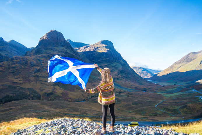 A woman waving a Scottish flag in the wind against the stunning backdrop of Glen Coe on a bright, sunny day with clear blue skies.