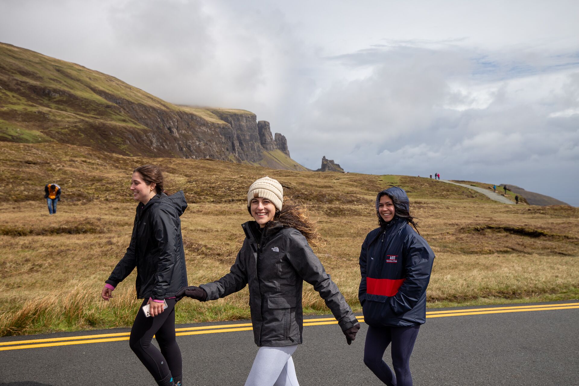 Three young women walking along a road on the Isle of Skye with the dramatic Quiraing hills in the background